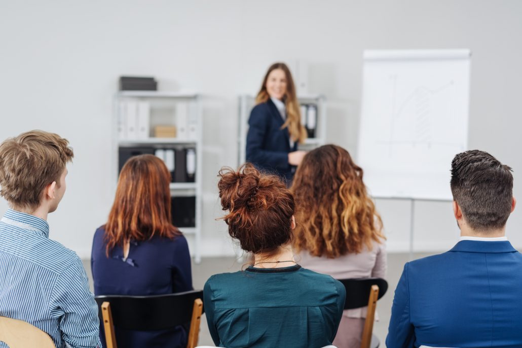 Group of young professional people in a meeting or training session sitting listening to a businesswoman give a presentation viewed from the rear