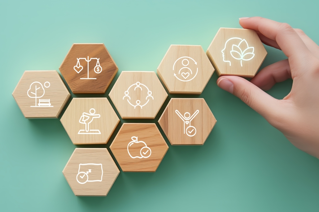 Die Erhaltung und Verbesserung der Gesundheit aller Mitarbeitenden ist zentrales Anliegen des Gesundheitsmanagements der Universität Klagenfurt. 2017 wurde sie mit dem Preis für Betriebliche Gesundheitsförderung ausgezeichnet. | Foto: Adobestock/1539859807/Paradee/KI-generiert Hellbraune hexagonale Holzwürfel mit Gesundheitssymbolen auf pastellgrünem Grund. Eine Hand greift nach dem Würfel in der Ecke rechts oben.