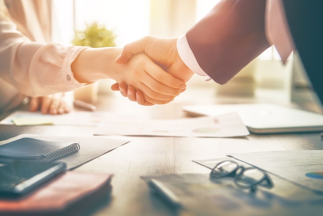 Zwei Personen im Business-Outfit schütteln sich über einem Schreibtisch mit Schreibutensilien die Hand.