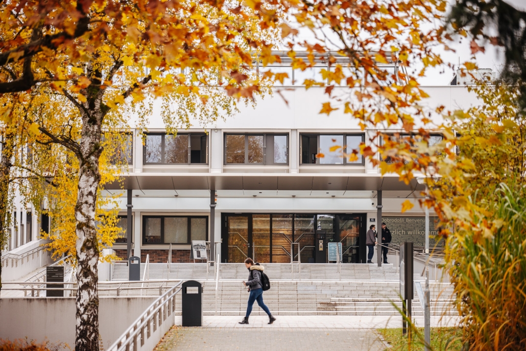 Person geht vor der Treppe des Eingangsbereichs zum Zentralgebäude der Universität Klagenfurt vorbei. im Vordergrund sind Bäume in herbstlichen Farben zu sehen.