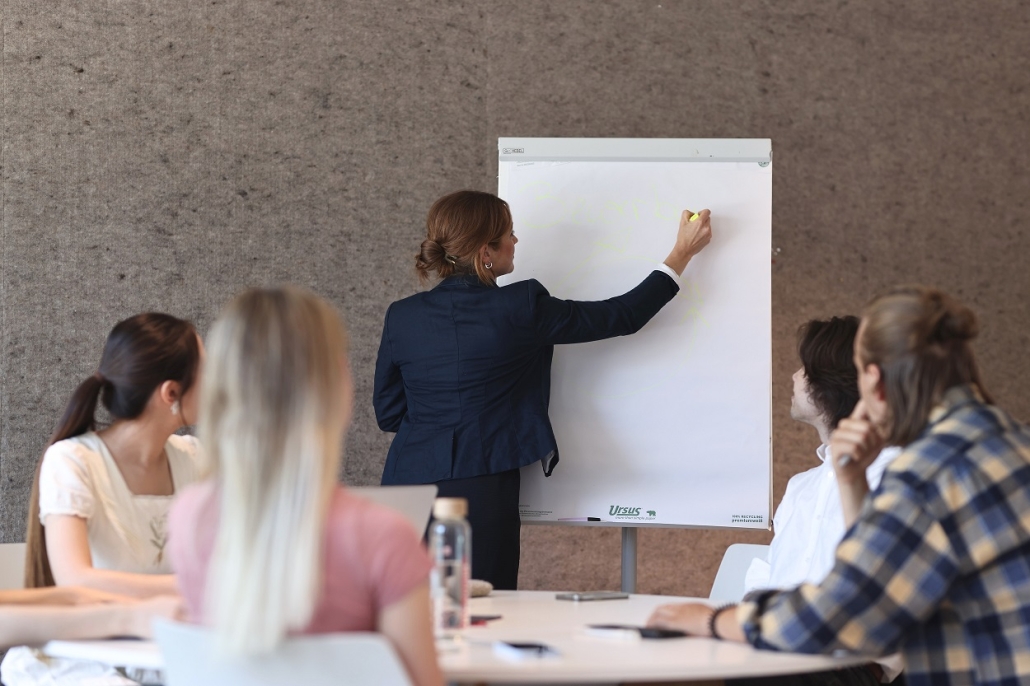Vier Personen sitzen an einem runden Tisch im Vordergrund und wenden ihren Blick zu einer Lehrperson, die auf einem Flipchart schreibt.