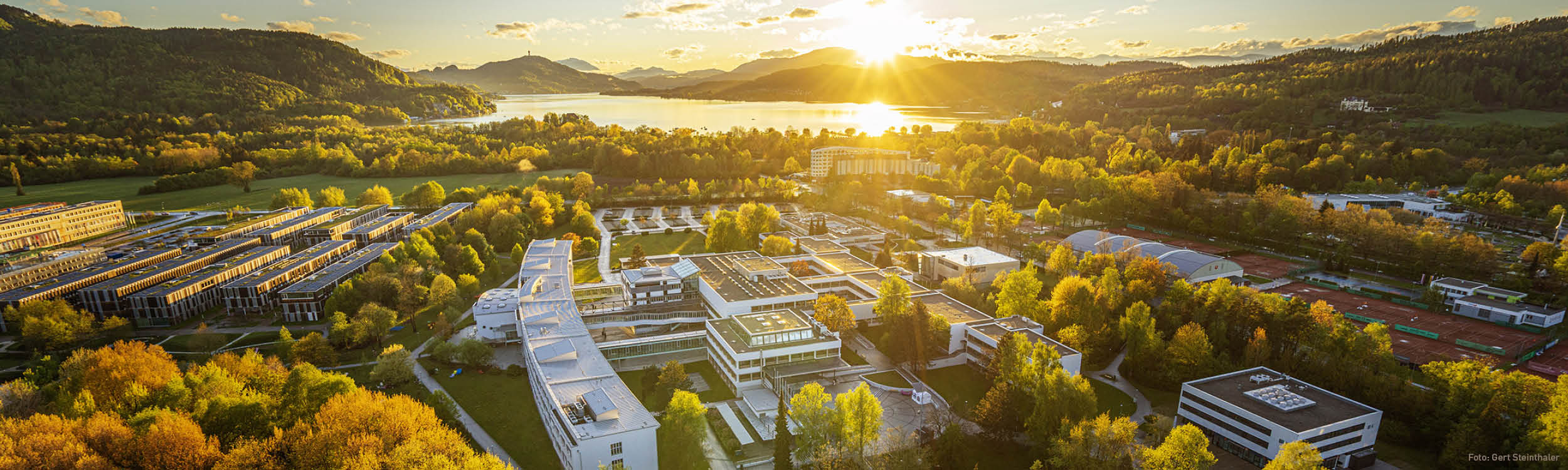 Im Vordergrund des Luftbildes sieht man das Gelände der Universität Klagenfurt und des Lakeside Parks, im Hintergrund steht die Sonne tief über dem Wörthersee und färbt die Landschaft in goldenes Licht.